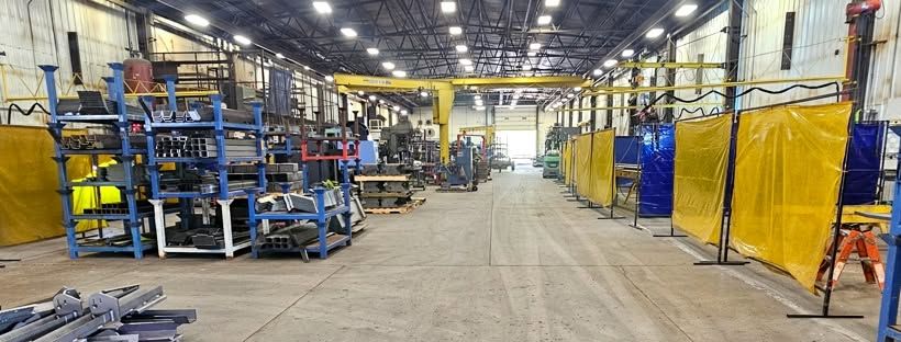 Shop floor with racks of steel stock and formed parts beside a row of welding bays with yellow and blue curtains