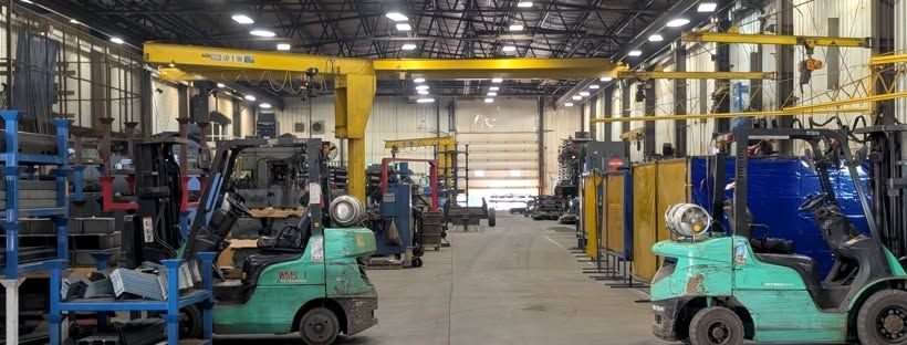 Shop floor with two forklifts, yellow jib and overhead cranes, welding bays, and a large overhead door