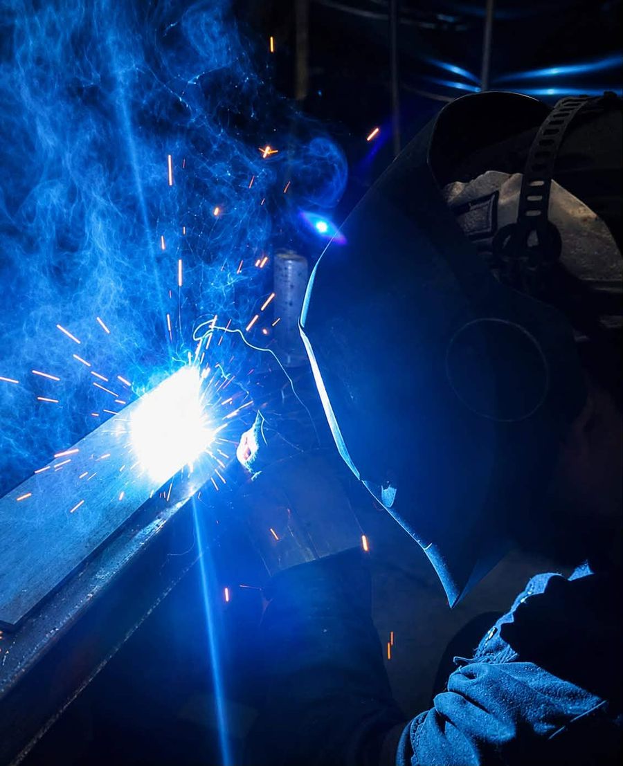 Close-up of a welder in a helmet welding a steel bar, with sparks and blue smoke